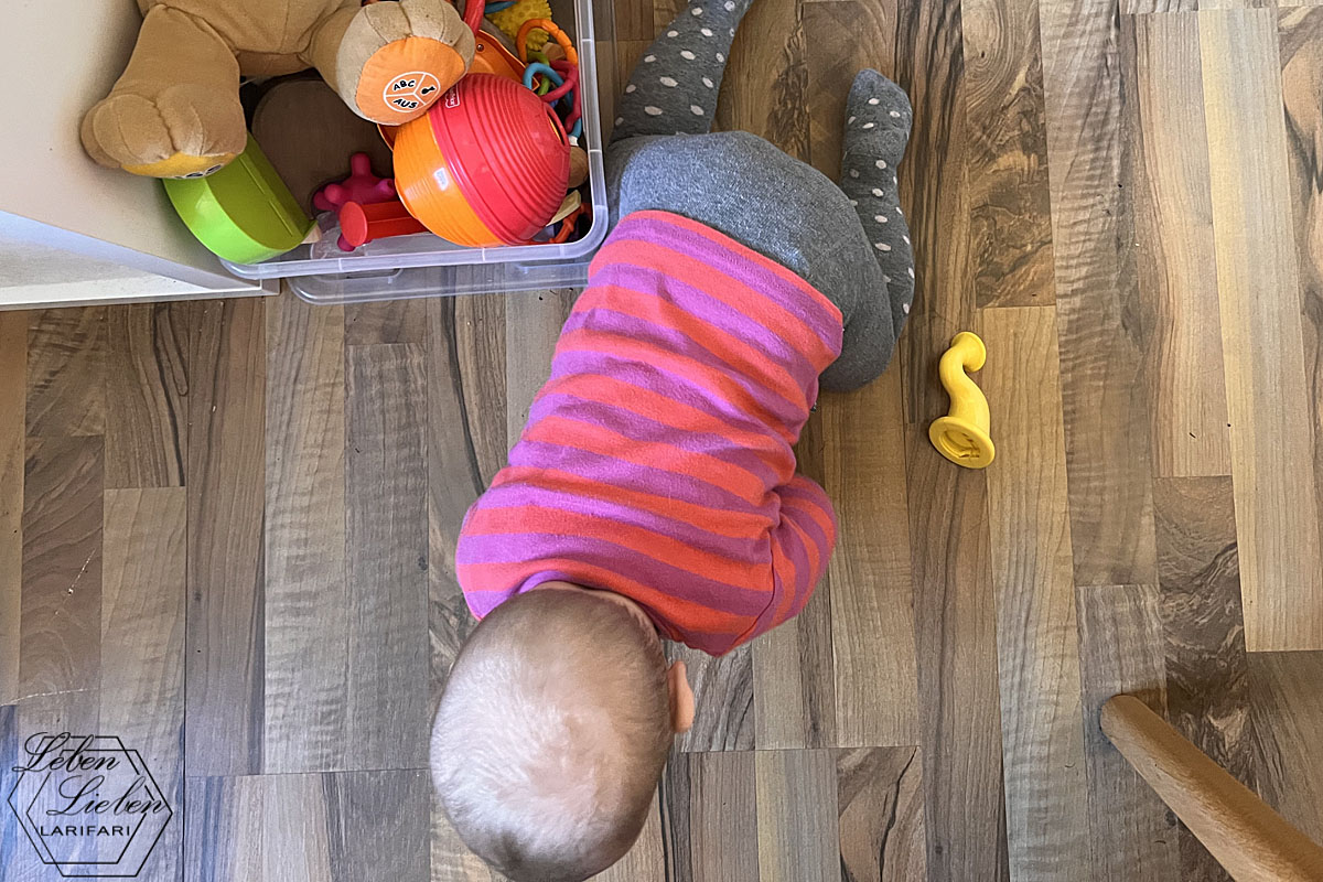 Ein Baby krabbelt über den Boden. Links steht eine Spielzeugkiste, rechts liegt eine Tröte.