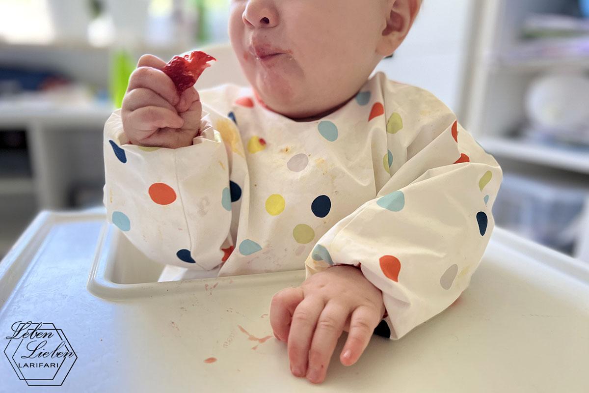 Baby sitzt im Hochstuhl, trägt ein gepunktetes Lätzchen und hat eine Erdbeere in der Hand.