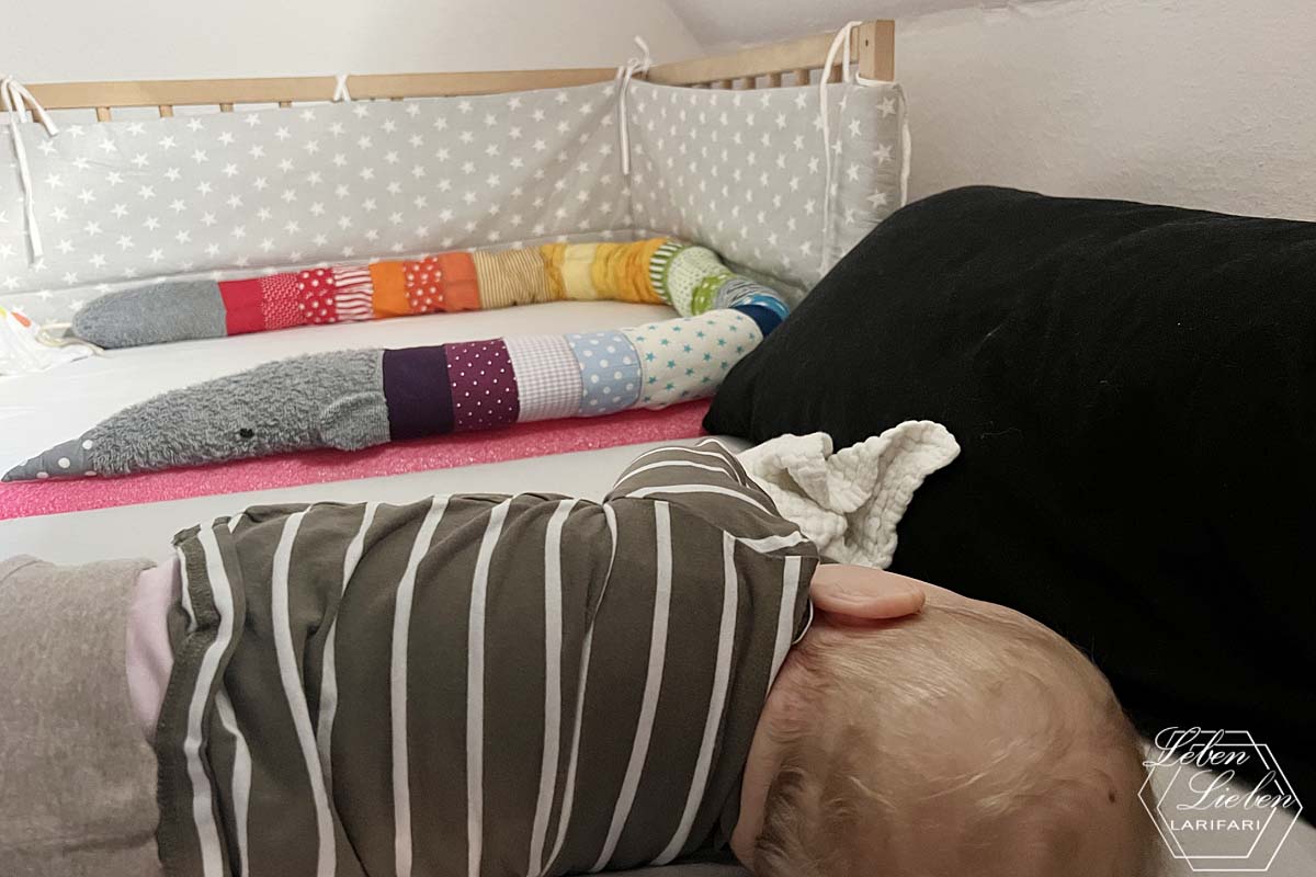 Baby liegt im Bett und schläft auf der Seite. Im Hintergrund ist das Babybett zu sehen