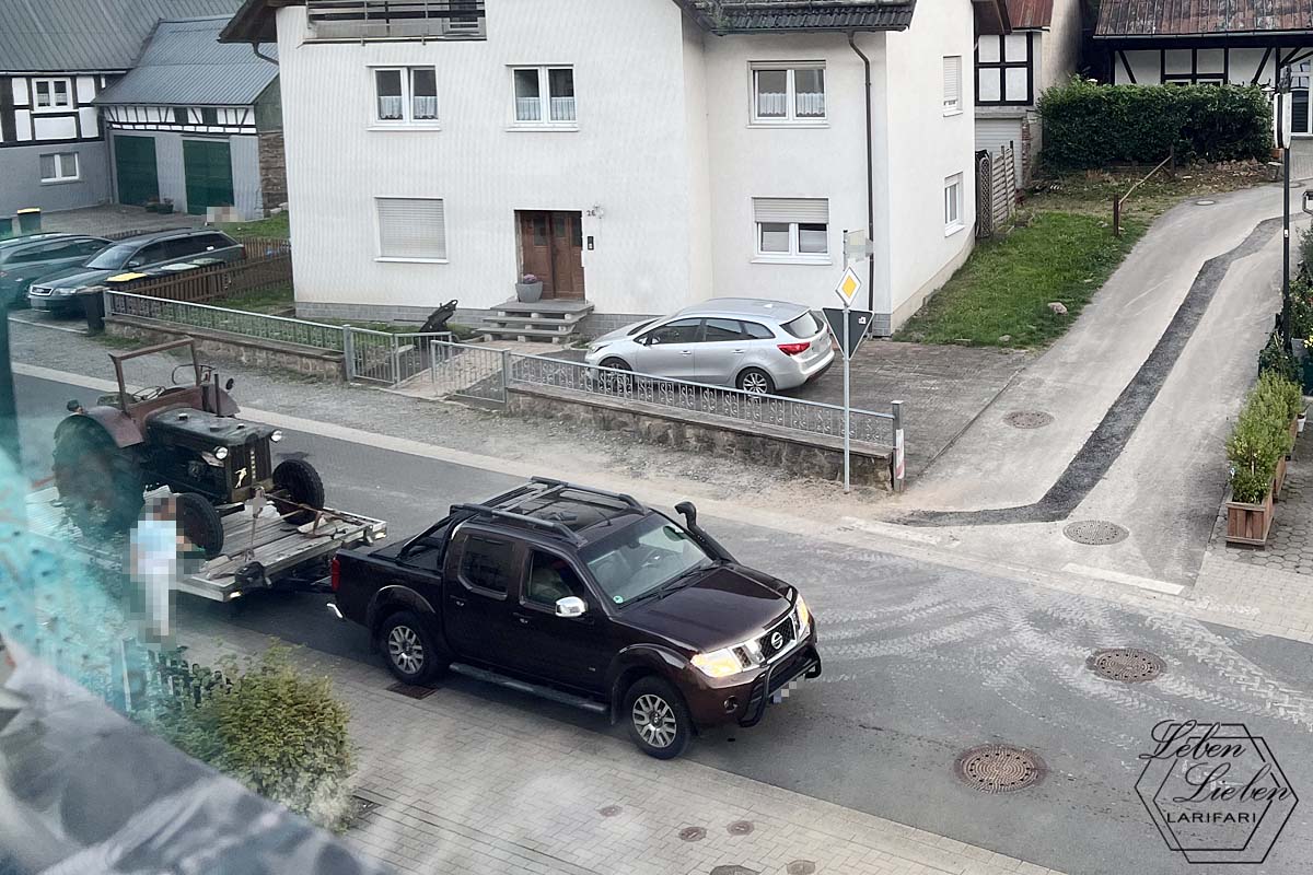 Eine Straße in einem Wohngebiet ist zu sehen. Ein Geländewagen hat auf einem Anhänger einen Trecker stehen