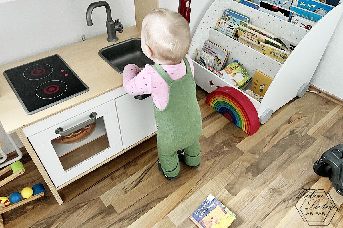 Ein Baby steht vor der Kinderküche, dahinter ist ein Bücherregal zu sehen
