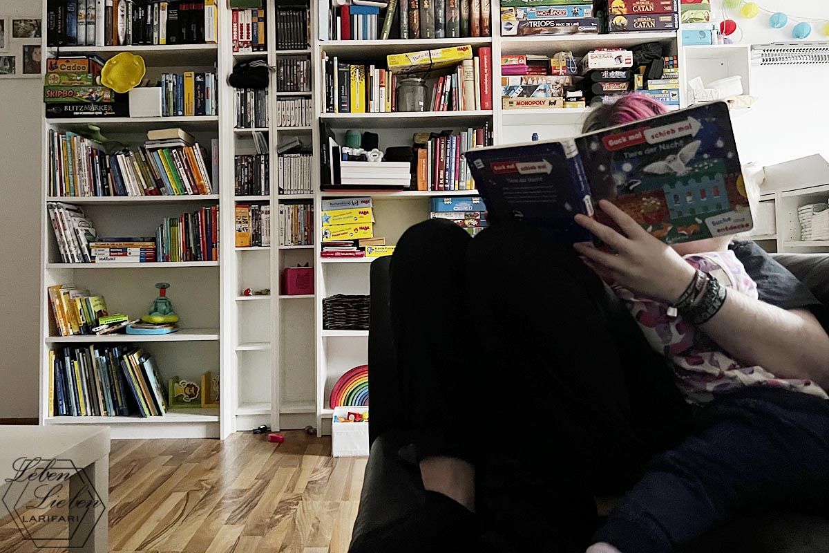 Eine junge Erwachsene hat ein Kleinkind im Arm, gfemeinsam sehen sie sich ein Bilderbuch an. Im Hintergrund steht ein chaotisches Bücherregal.