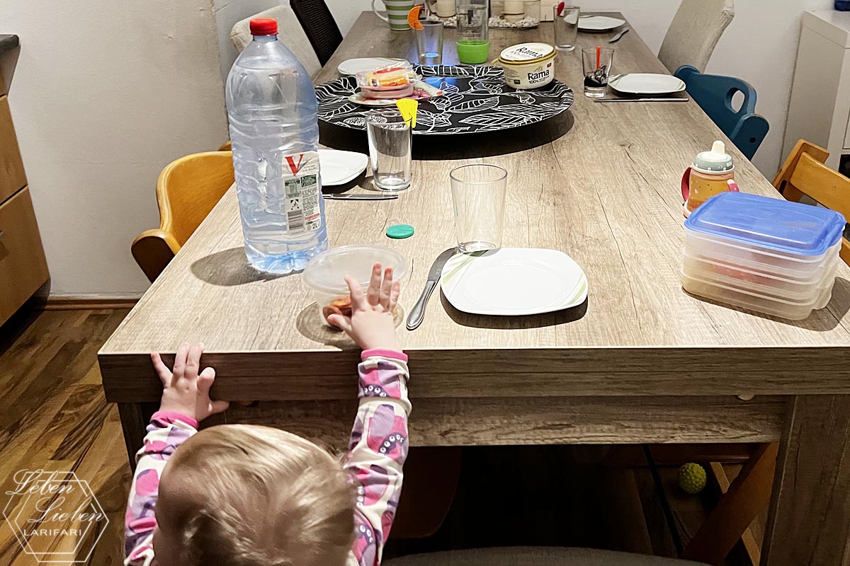 Es ist ein langer, gedeckter Tisch zu sehen, an dessen Kopfende ein Kleinkind etwas auf den Tisch stellt.