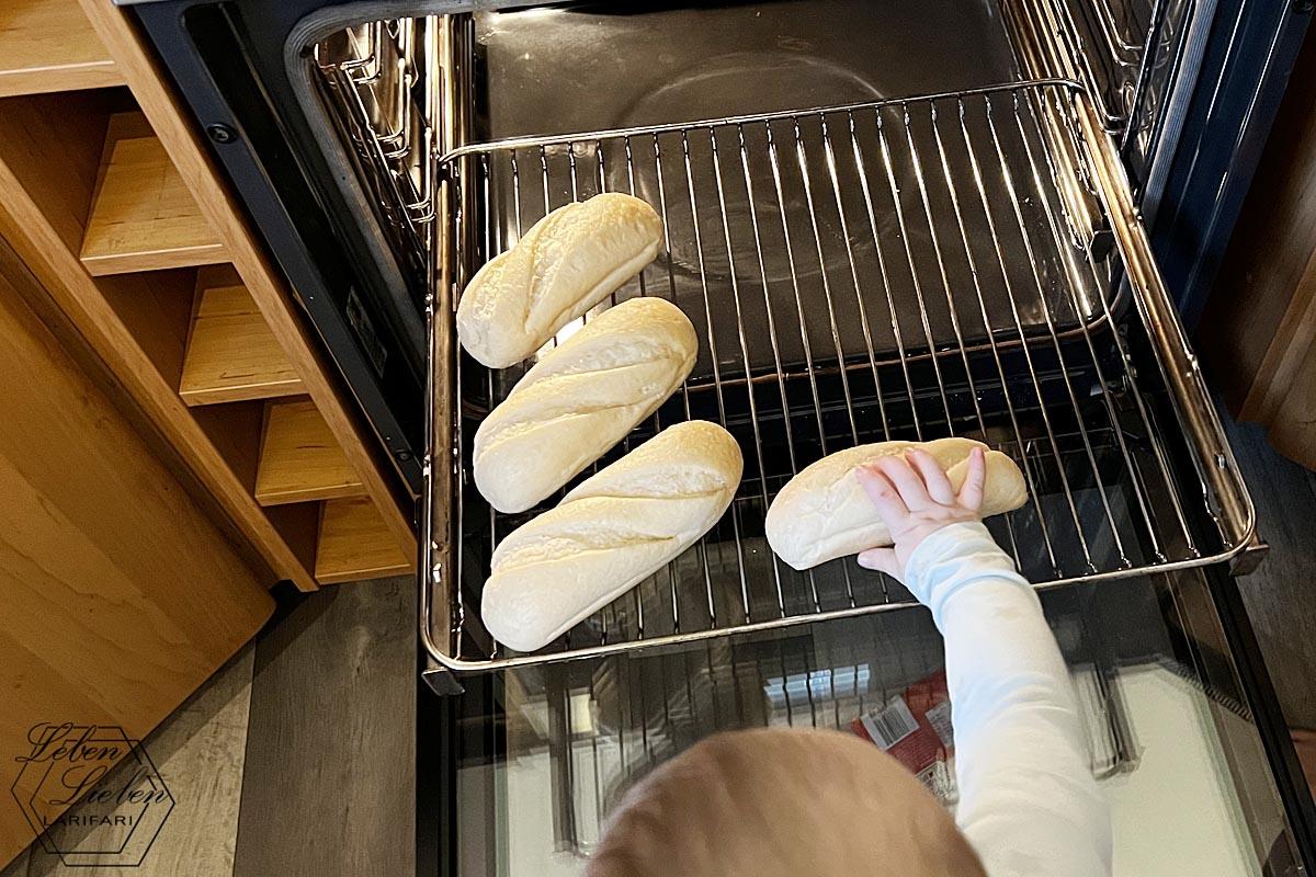 Vor einem offenen Ofen steht ein Kleinkind und legt Brötchen auf das Rost