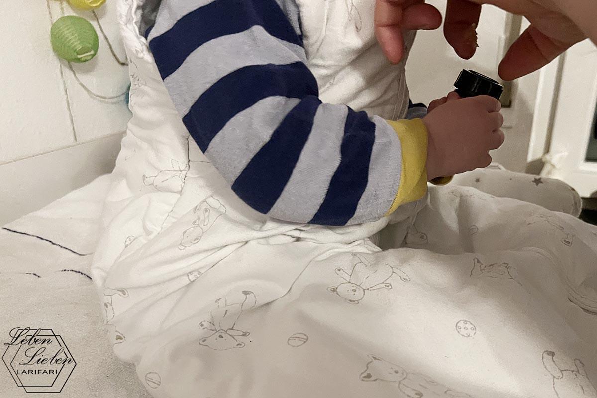 Ein in Schlafanzug und Schlafsack gekleidetes Kleinkind sitzt auf dem Wickeltisch und hat ein Döschen in der Hand