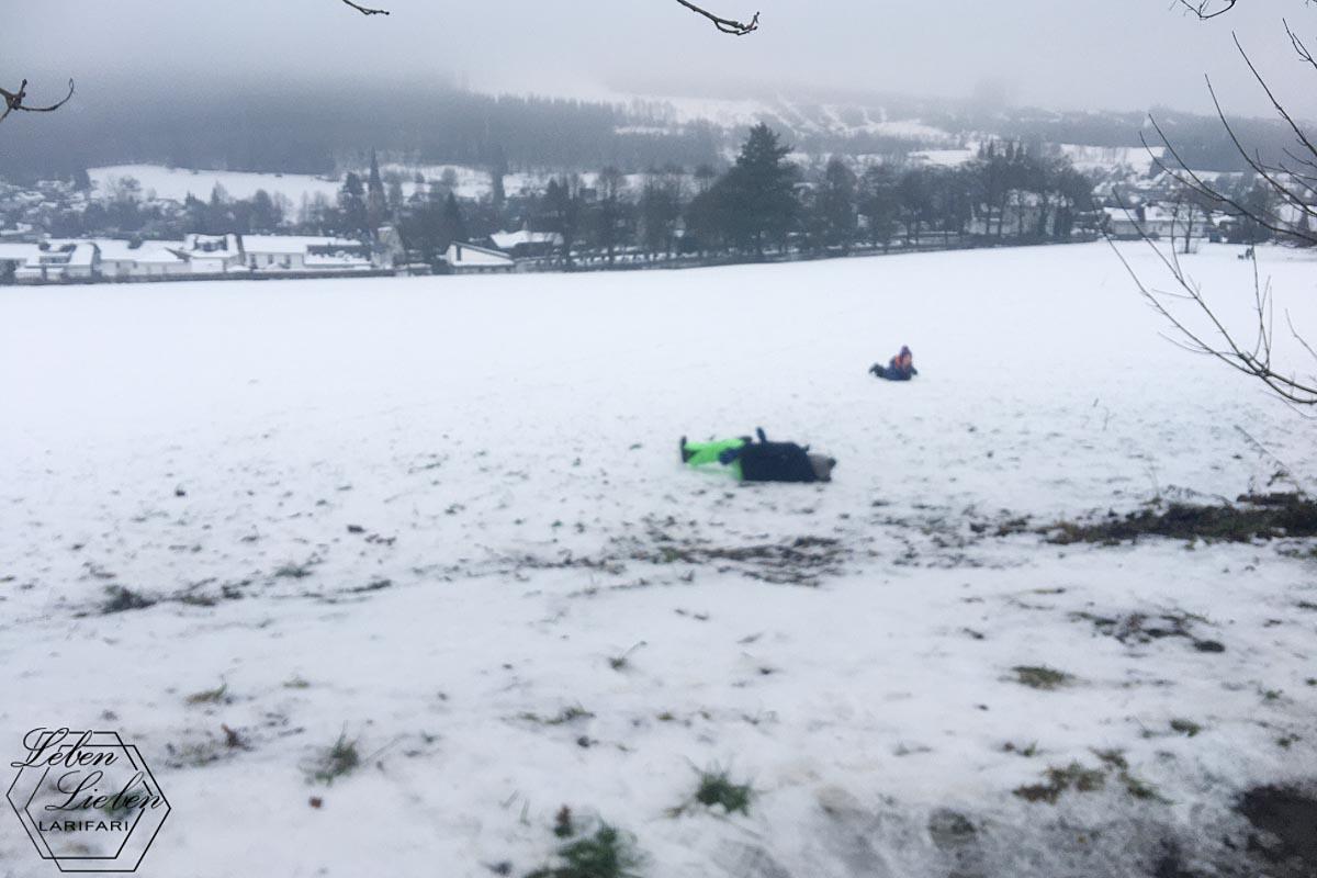 2 Kinder rollen eine verschneite Wiese herunter. Im Hintergrund ist das Dorf zu sehen-