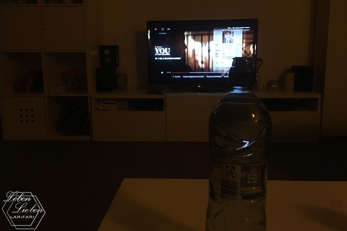 Eine Wasserflasche steht im Vordergrund auf einem Tisch. Im Hintergrund ist ein laufender Fernseher mit "YOU" zu sehen