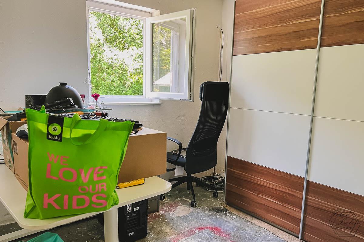 Ein Büro mit Kartons und Kram auf dem Schreibtisch, einem Schreibtischstuhl und einem Kleiderschrank auf Betonboden; im Hintergrund ein geöffnetes Fenster