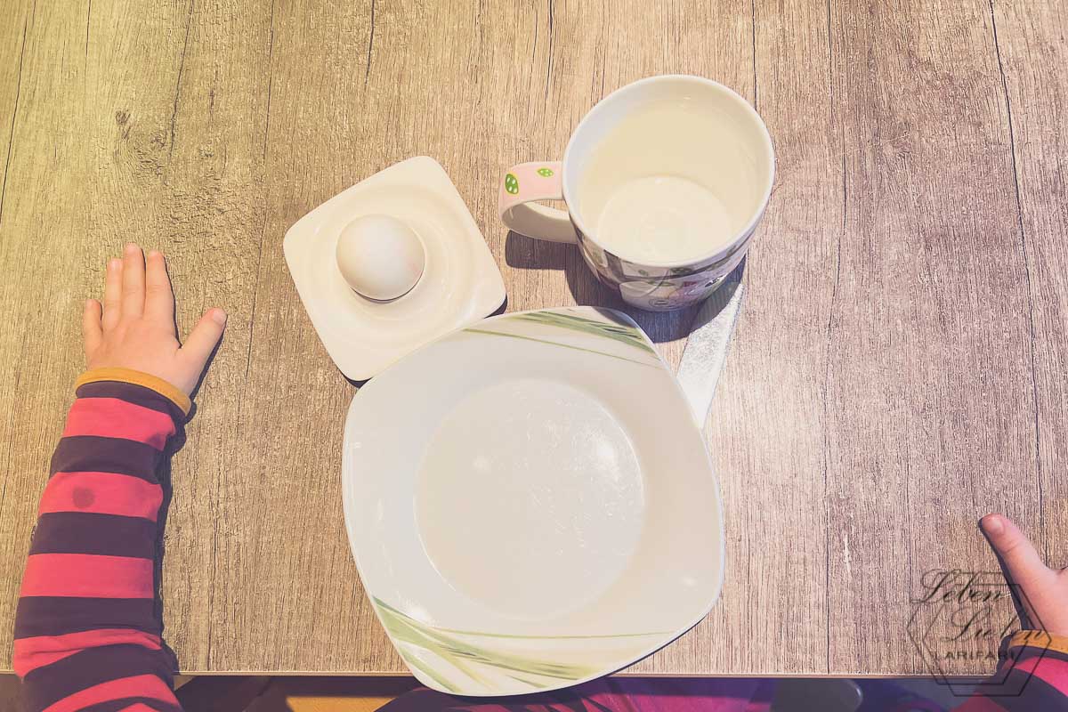Eingedeckter Platz mit Teller, Tasse und Eierschale am Tisch, zu sehen ist, dass ein Kleinkind am Platz sitzt. Ansicht von oben.