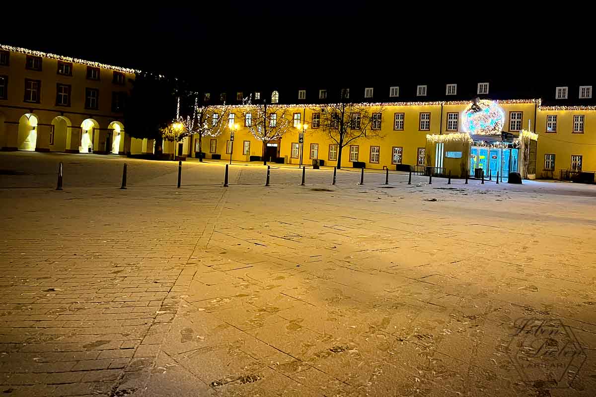 Ein Schlossplatz, leicht mit Schnee bedeckt. Das Gebäude ist noch weihnachtlich von Lichterketten erleuchtet.