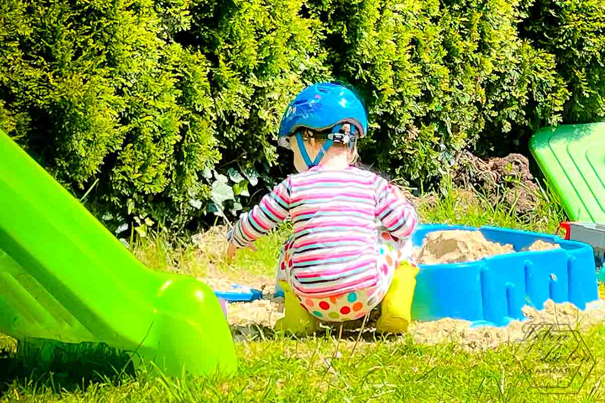 In der Bildmitte ist ein vor dem Sandkasten hockendes Kind zu sehen. Links im Bild ein Teil einer grünen Kinderrutsche.